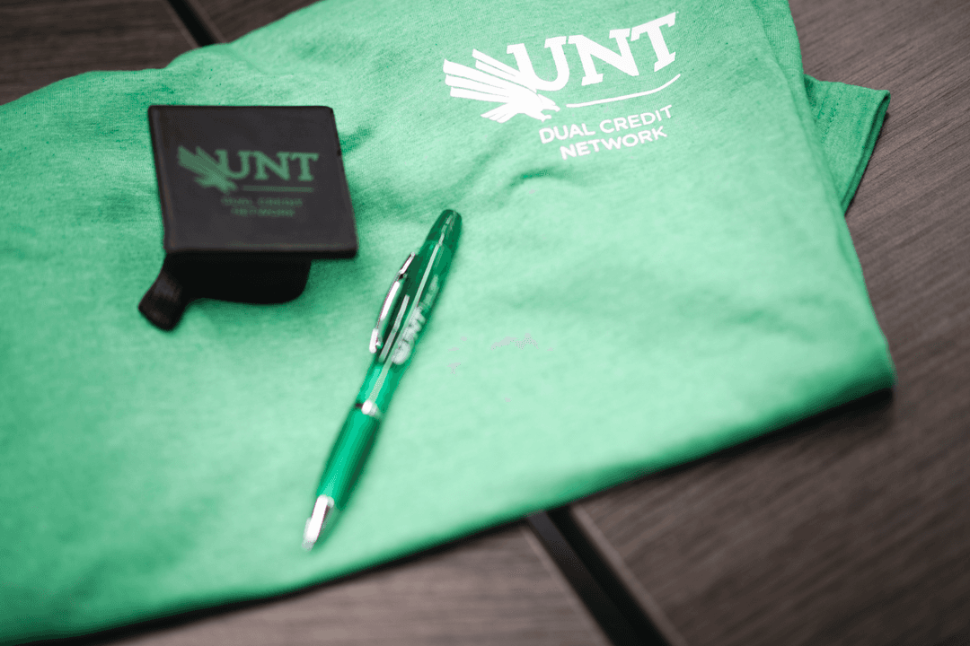 UNT Dual Credit Network green t-shirt, branded pen, and black box displayed on a wooden surface.