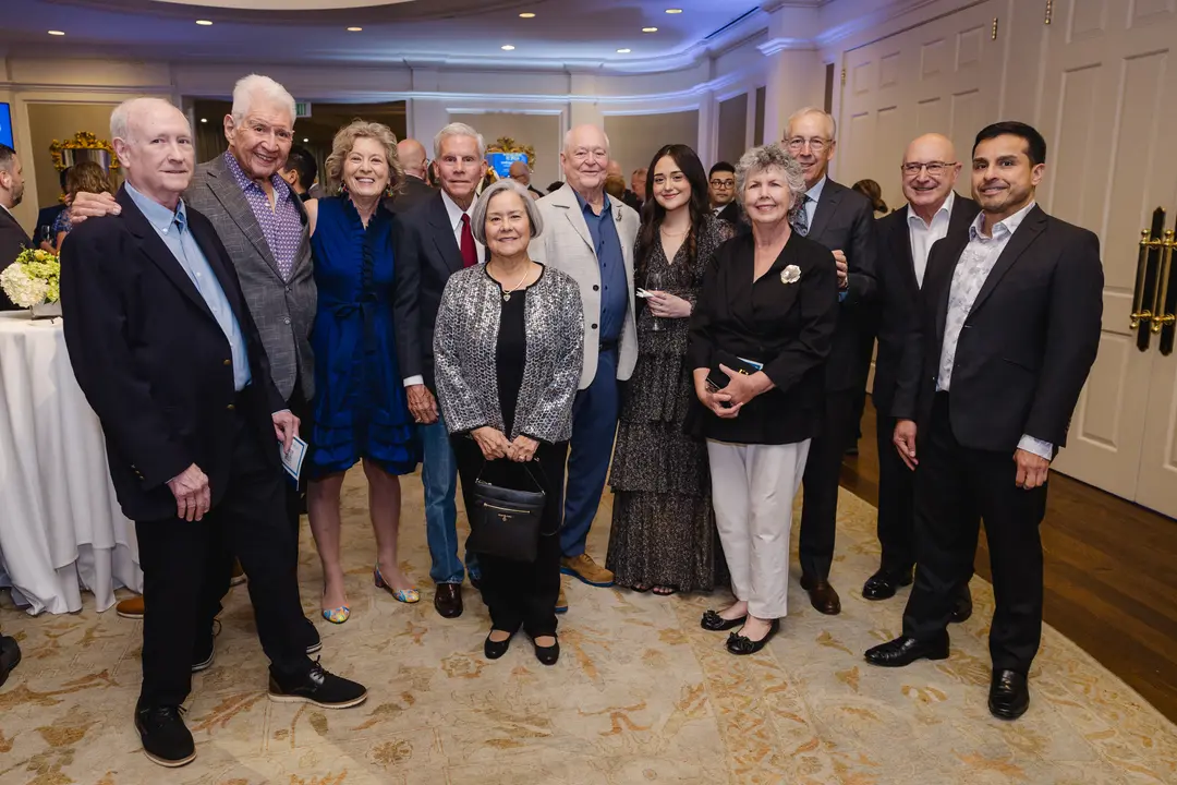 Group of eleven people in formal attire posing together at an indoor event in an elegant ballroom.