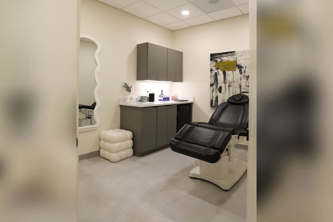 Modern aesthetic treatment room with black reclining chair, gray cabinets, wavy mirror and abstract wall art.