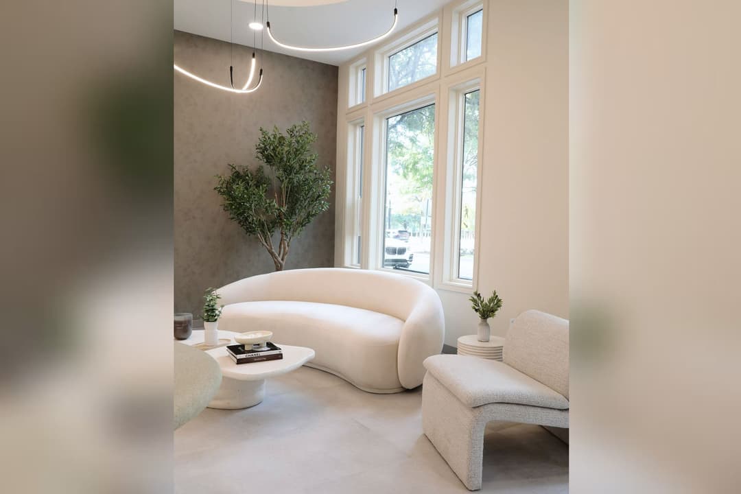 Modern lounge with cream curved sofa, armchair, coffee table with books, potted tree, and large windows.