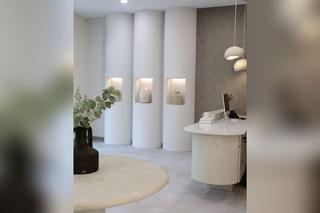 Minimalist beige reception area with curved columns, lit niches holding vases, a plant, and a curved desk.