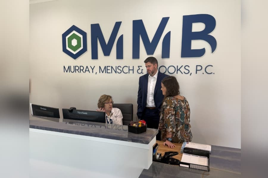 Three people converse at a reception desk beneath a Murray, Mensch & Brooks, P.C. "MMB" logo wall.