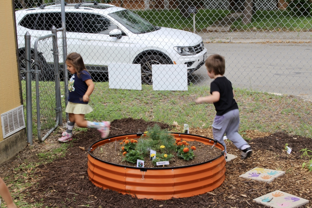Two children jump from tile to tile beside the garden bed, enjoying outdoor play near the plants they’ve helped tend (Jamie Moore/Community Impact)