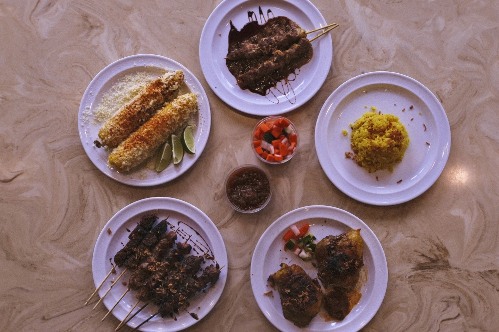 An assortment of Indonesian dishes from Sate Texas is displayed on a table, featuring grilled satay skewers, seasoned rice, corn topped with cheese and spices, and house-made sauces. (Manuel Holguin JR/ Community Impact)